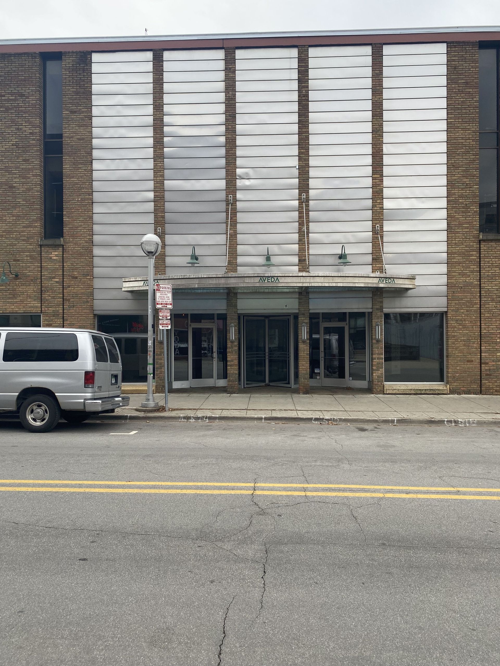 Exterior of a brown brick building with a metal facade and glass doors, parked van in front.