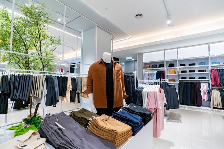 Bright clothing store interior with racks of folded pants, hanging clothes, and a mannequin in a brown jacket