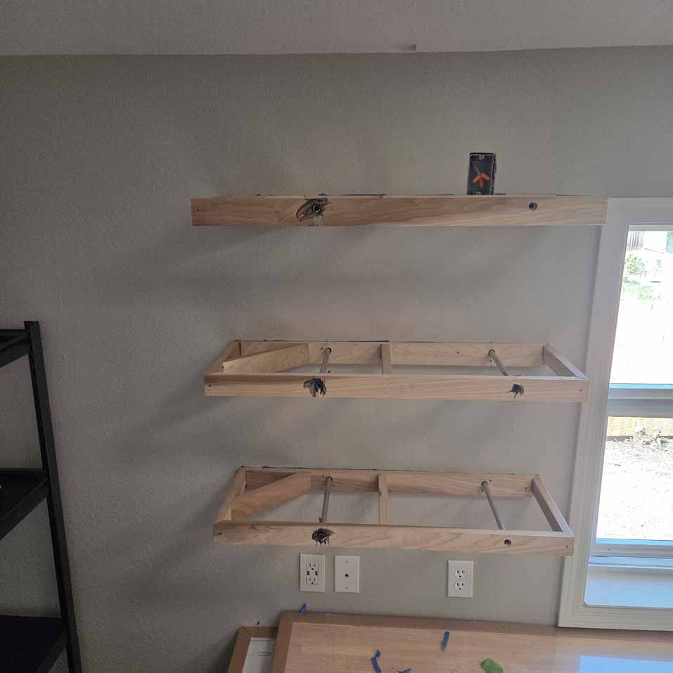 Three wooden wall shelves mounted above outlets beside a window, with a small can on the top shelf.