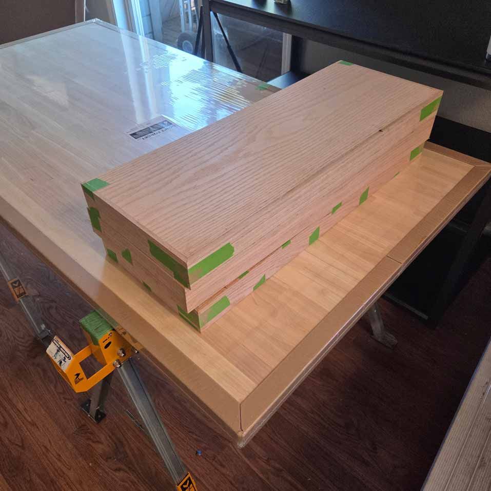 Stacked wood boards with green tape on a table, next to a yellow clamp in a workshop.