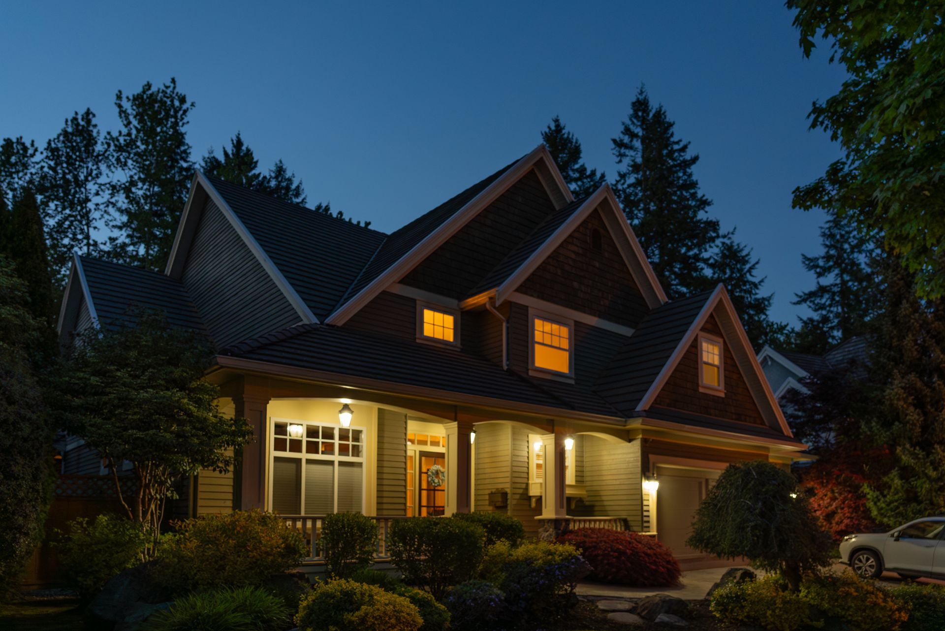 Warmly lit cabin-style house at dusk with a porch, dark roof, and trees surrounding it