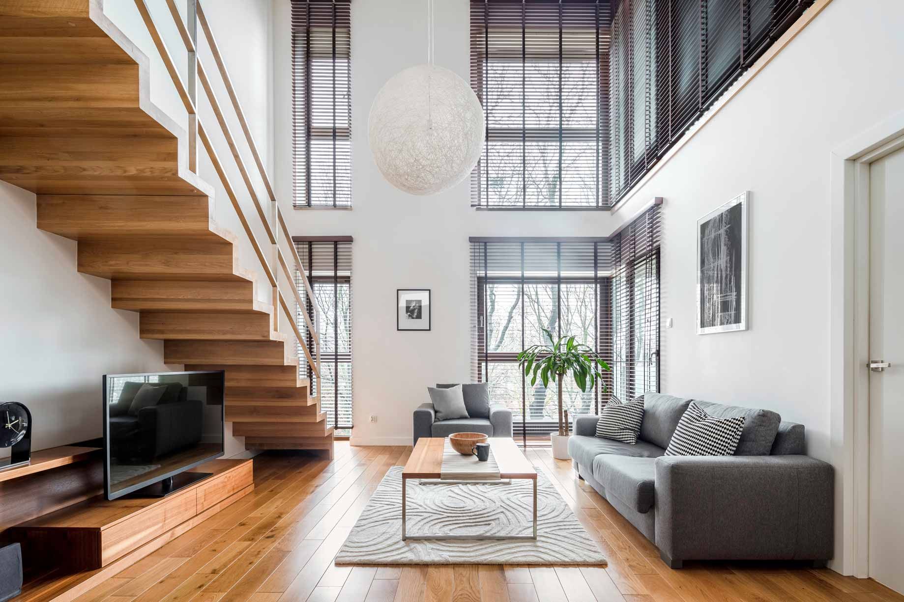 Modern loft living room with wooden staircase, gray sofa, TV, tall windows, and natural light