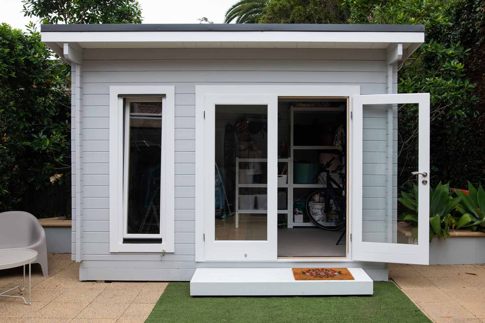 Small white backyard shed with open doors, front steps, and outdoor seating on either side