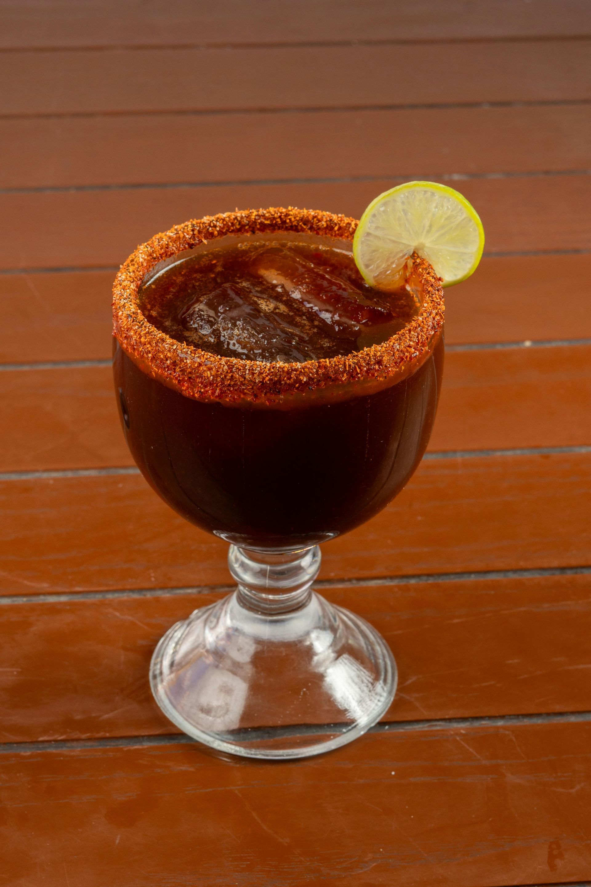 Brown cocktail in a goblet glass, rimmed with chili powder, garnished with a lemon slice.