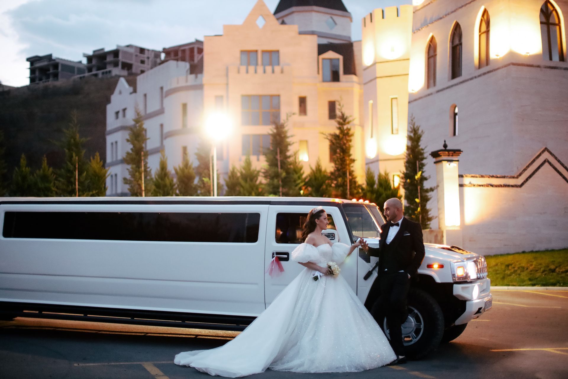 A bride and groom are standing next to a limousine in front of a castle.