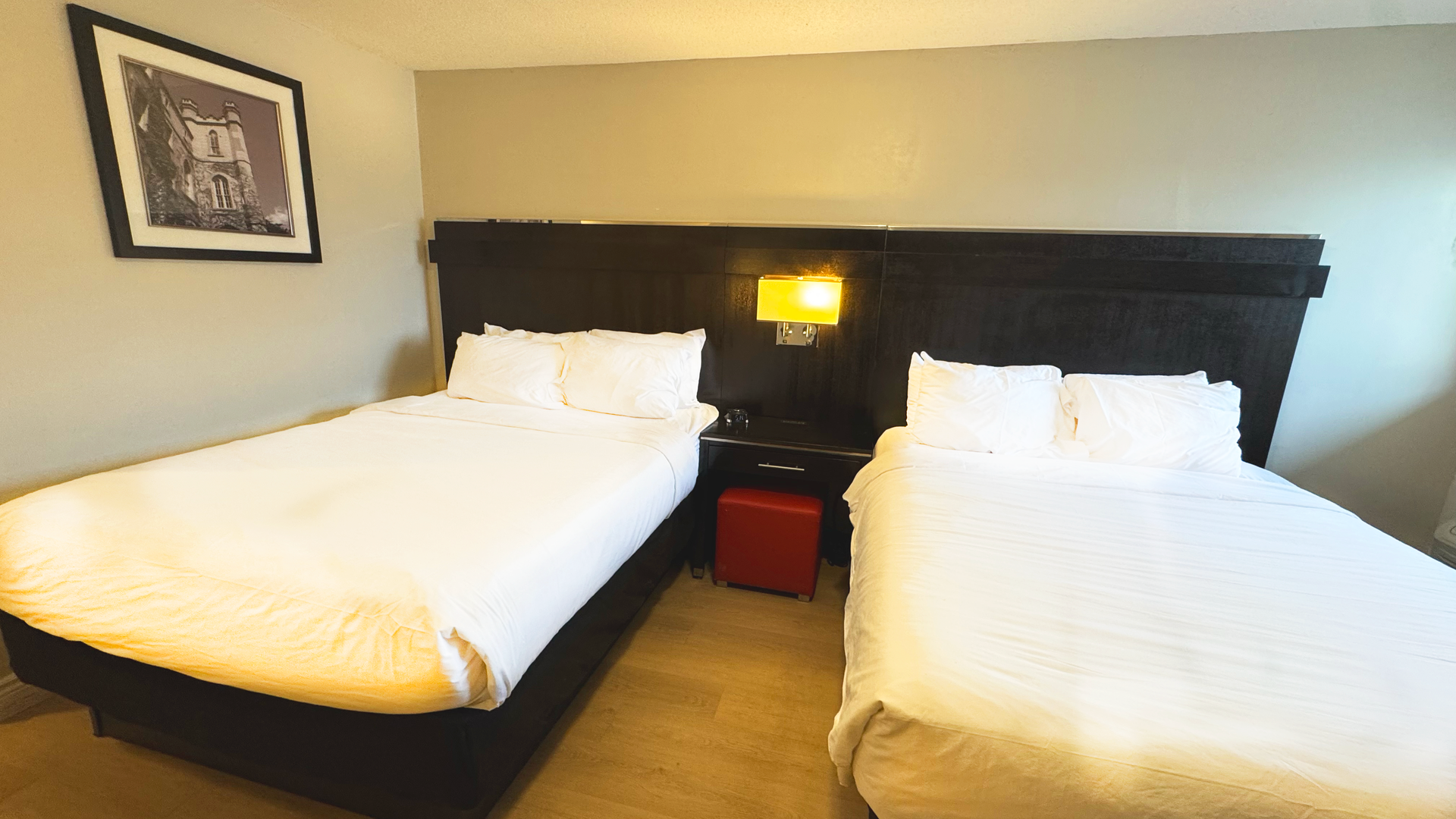 Hotel room with two beds, nightstand, and framed art. Beds have white linens, dark headboard.