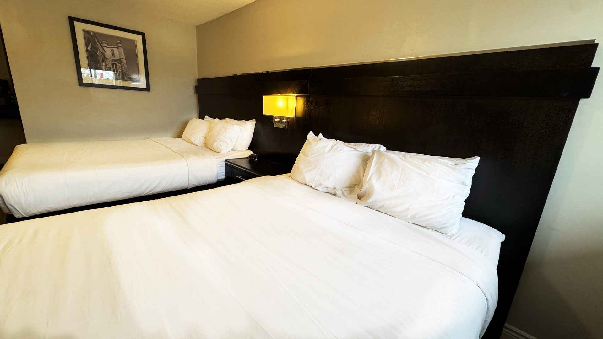 Two beds with white linens in a hotel room, dark headboard, small picture on the wall, and bedside lamp.