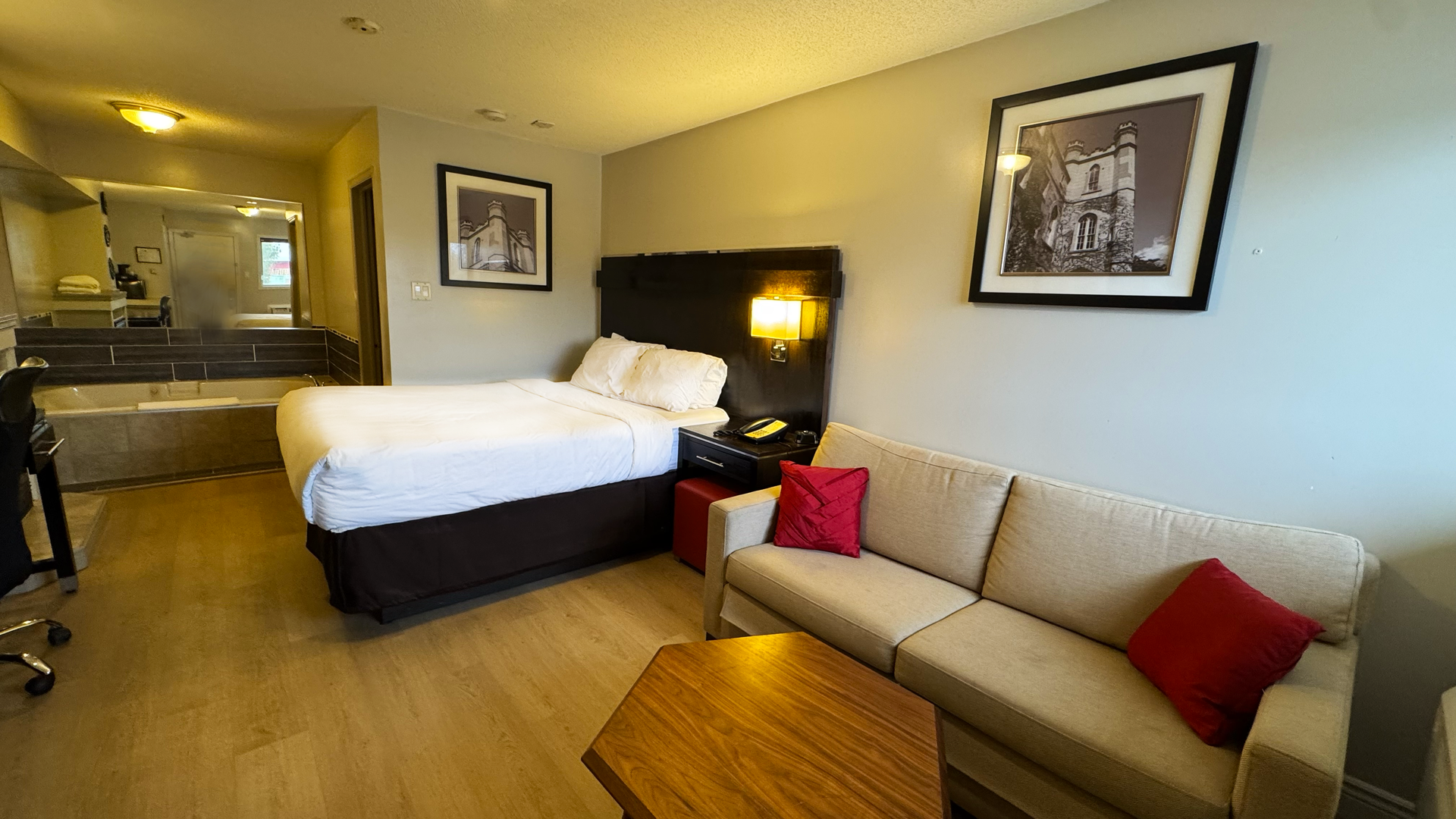 Hotel room with bed, sofa, desk, and a large Jacuzzi. Neutral tones with red accent pillows.