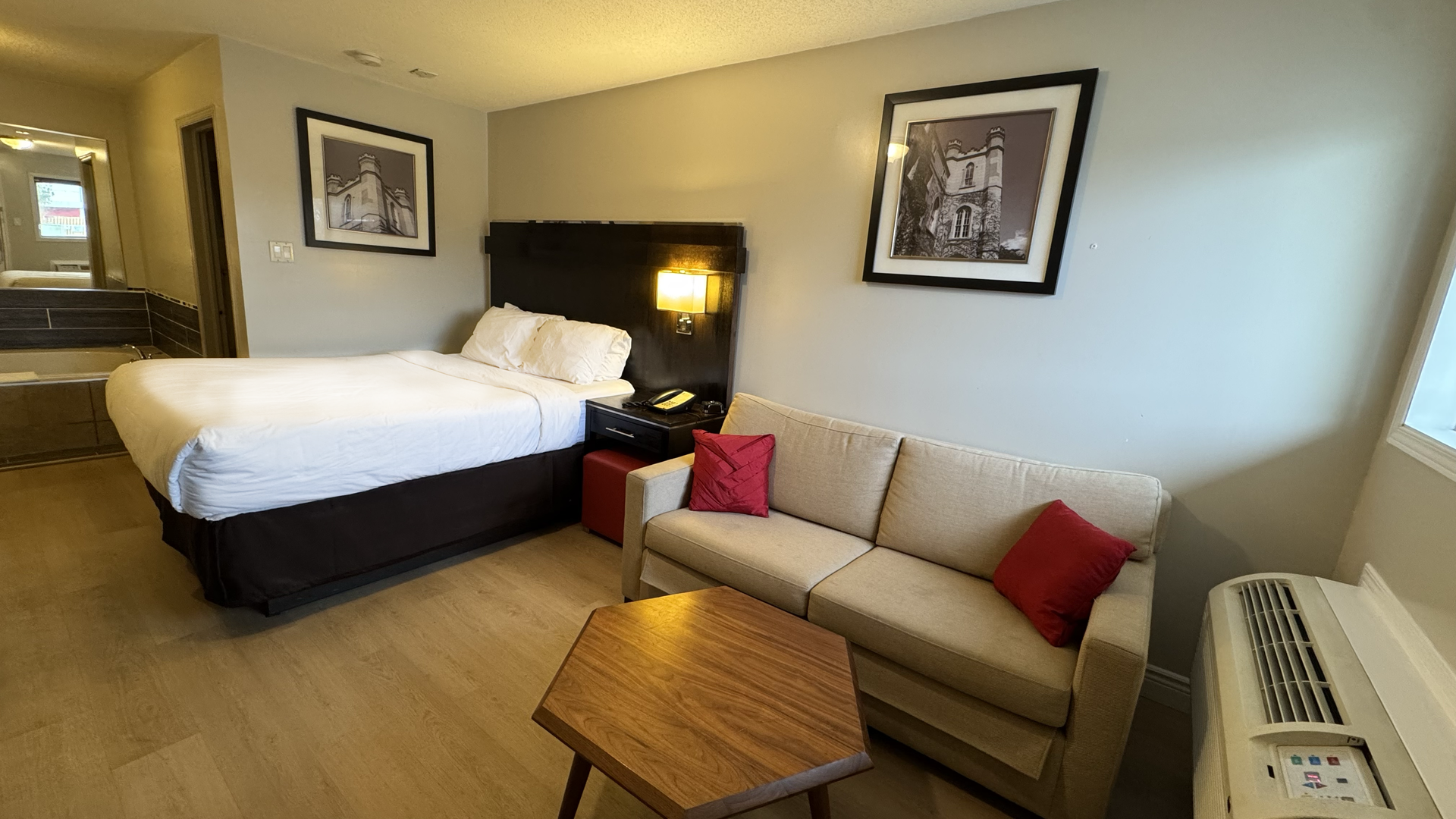Hotel room interior with a bed, sofa, side table, artwork, and window.