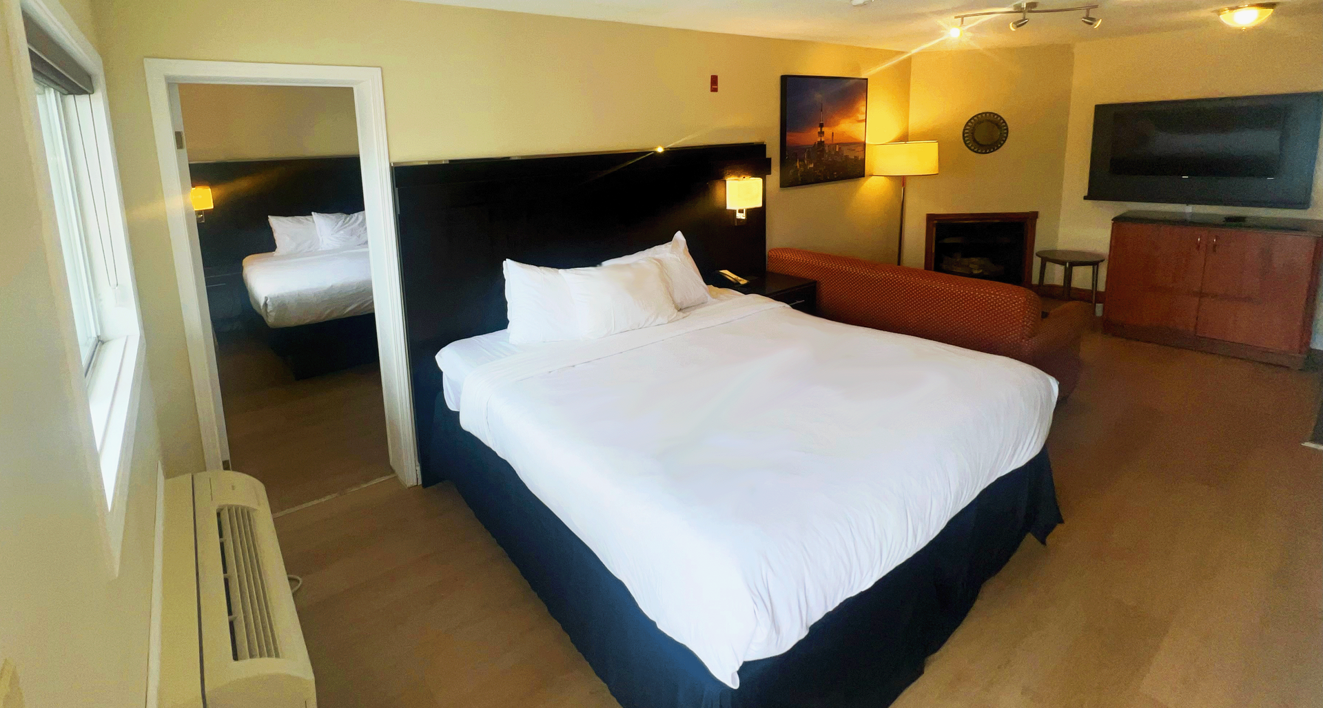 Hotel suite with a king-sized bed, fireplace, TV, and an open doorway to a second bed.