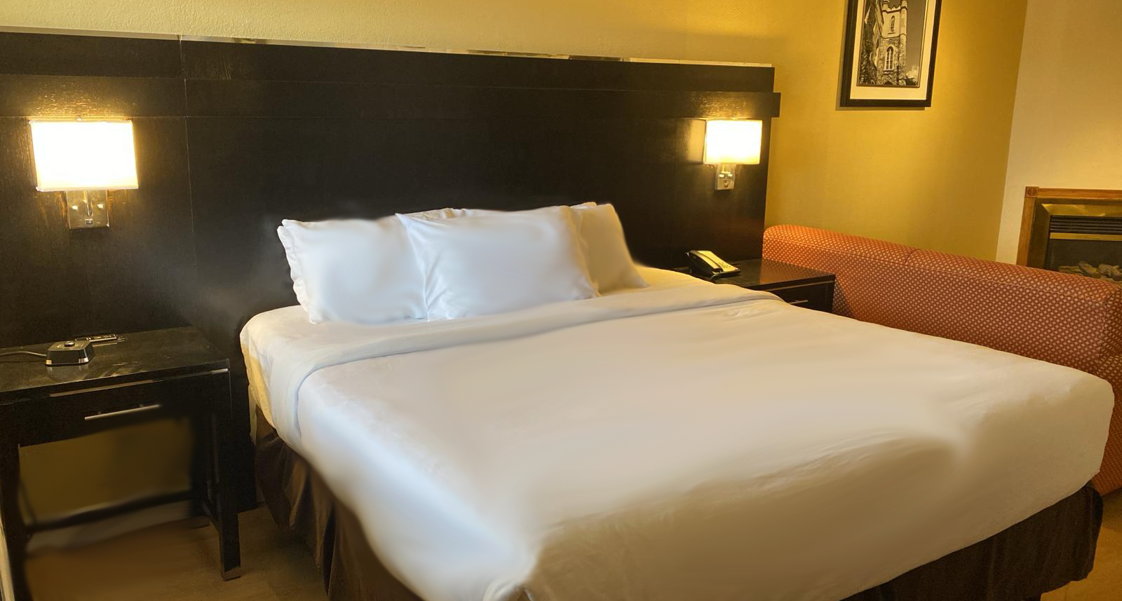 A hotel room with a made bed, nightstand, and small sofa. Soft lighting and neutral colors.