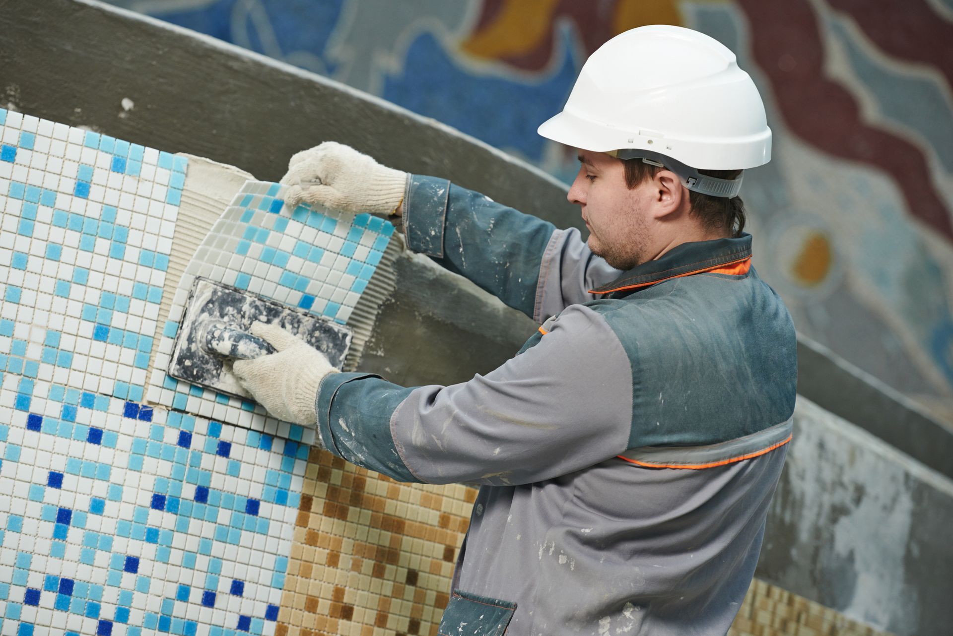 Professional tile setter installing blue mosaic tiles on wall wearing hard hat and work gloves.