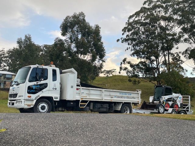 White truck with a trailer carrying a small Bobcat loader on a gravel road — Concrete Services in Coffs Harbour, NSW