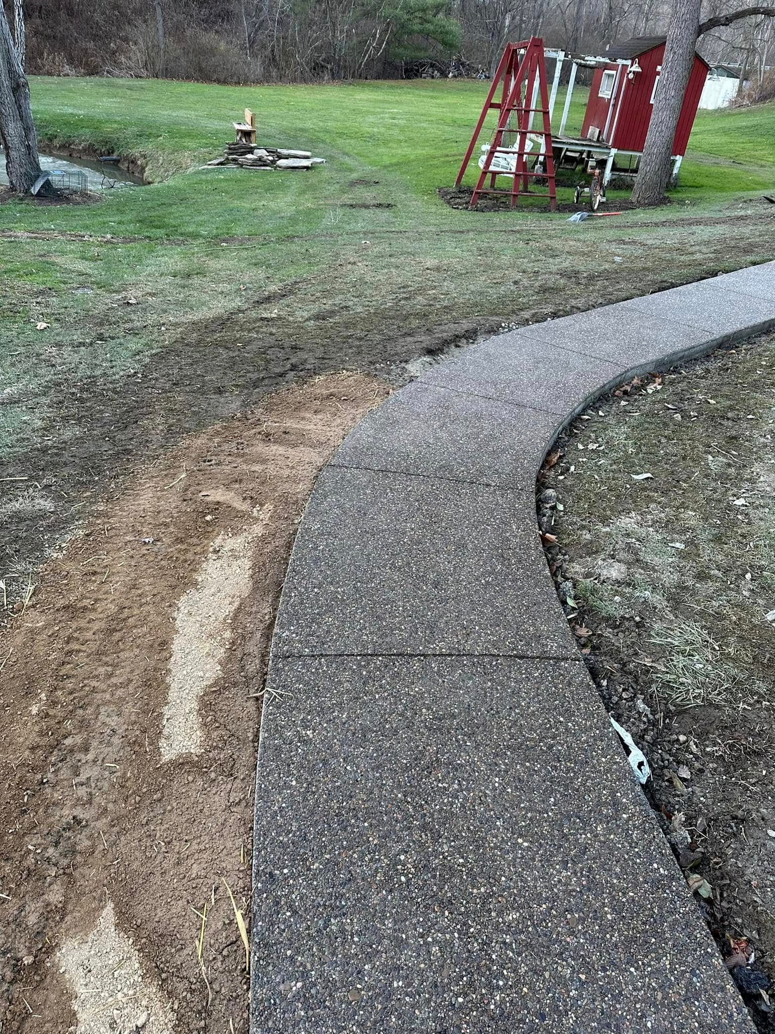 Curved concrete walkway beside eroded gravel edge in a grassy yard, with red outdoor equipment in the background