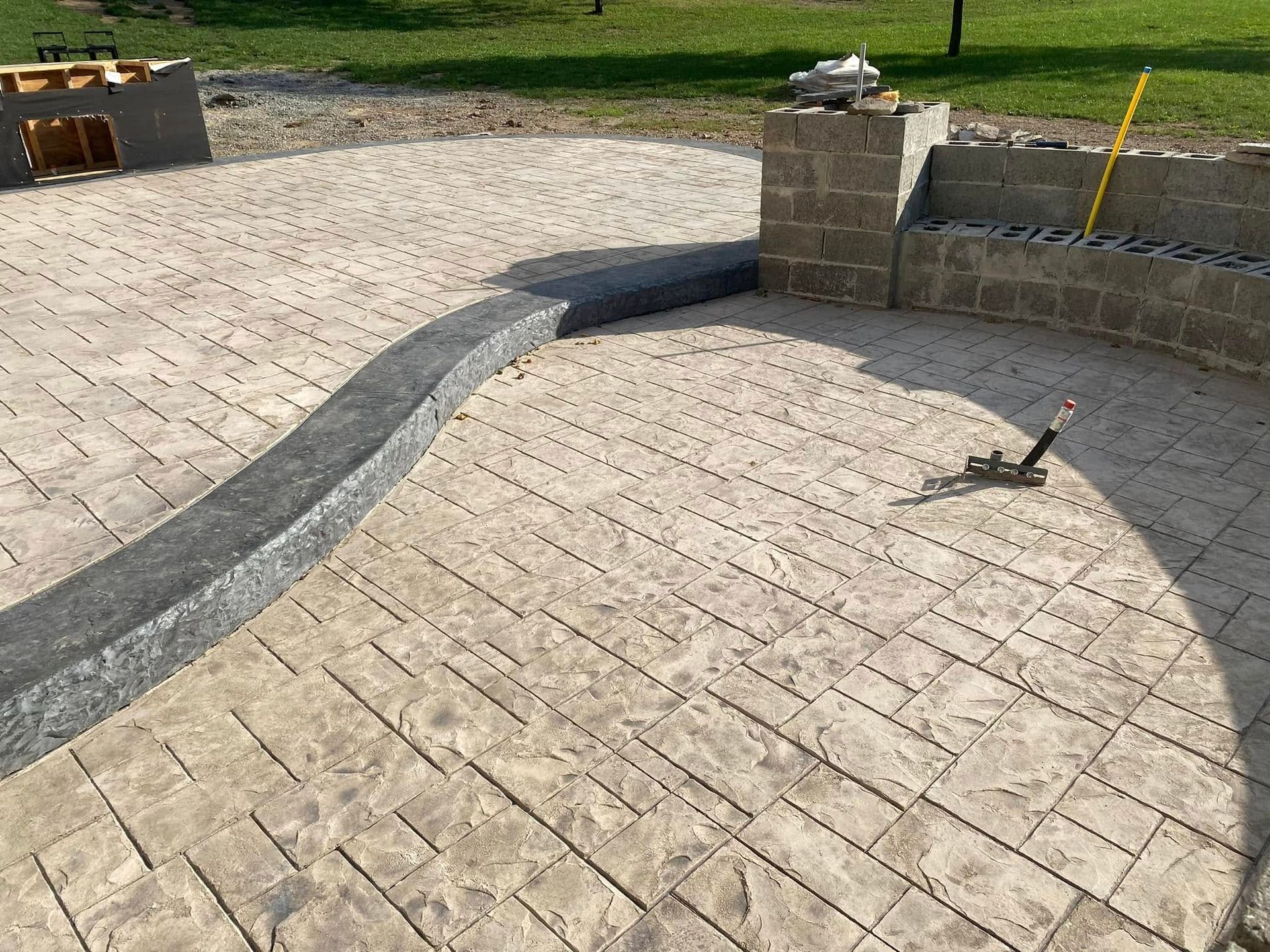 Paved patio with curved dark border beside a low stone wall and grassy yard