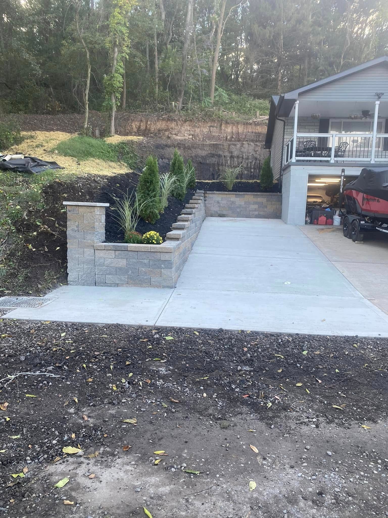 Concrete driveway beside a stone retaining wall and landscaped garden leading to a garage.