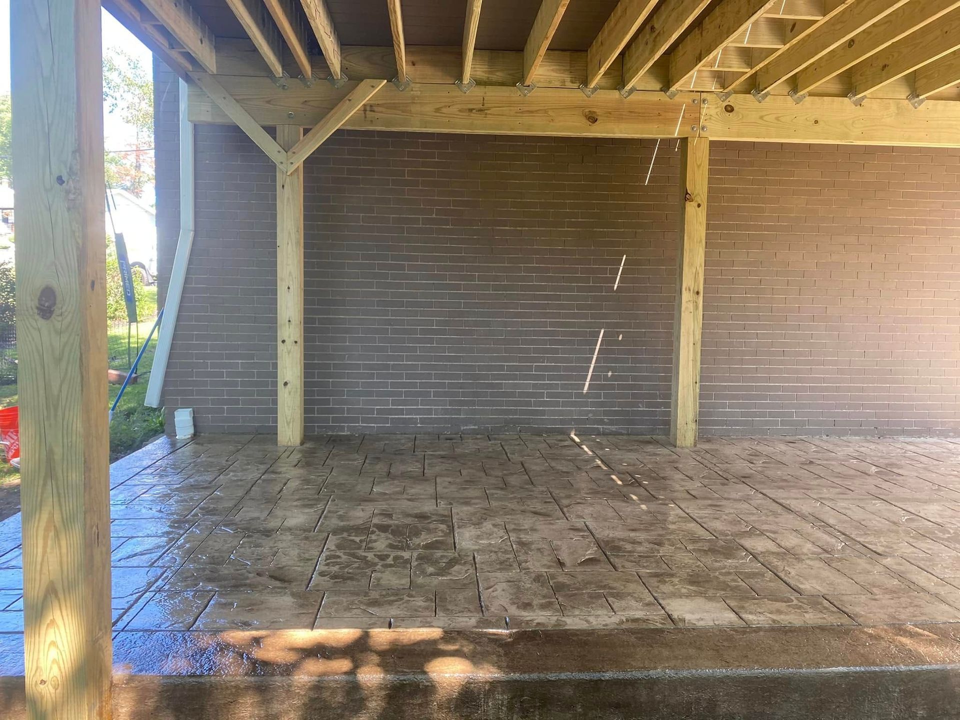Empty covered patio with stone floor, exposed beams, and brick walls, lit by sunlight.