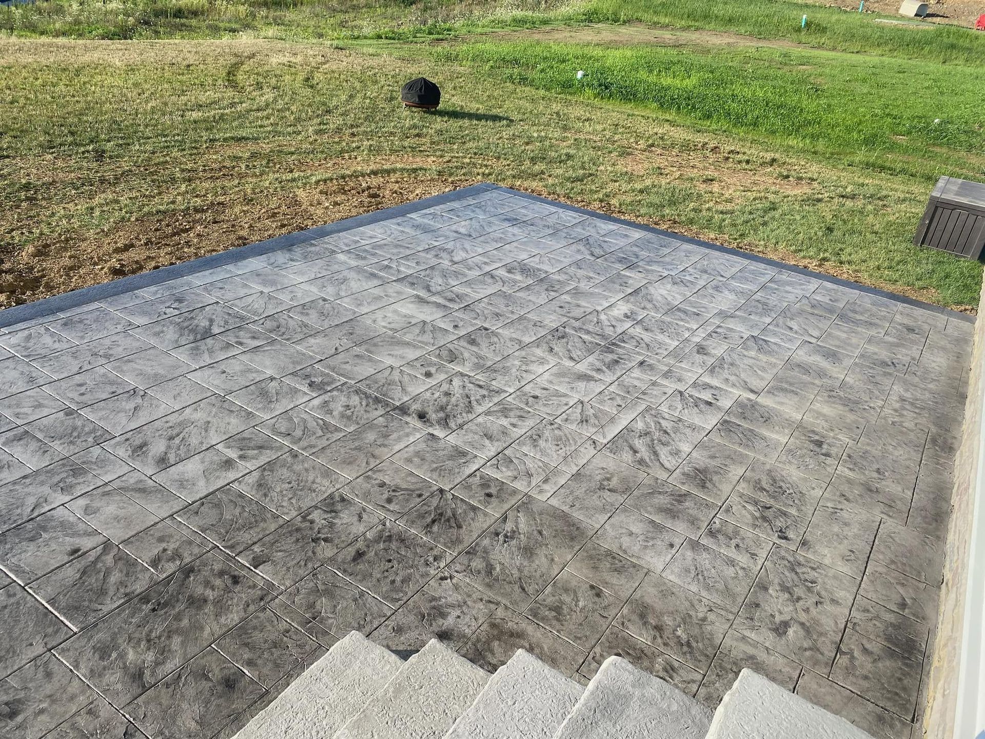 Gray paved patio in a grassy yard, with a small black dog in the background.