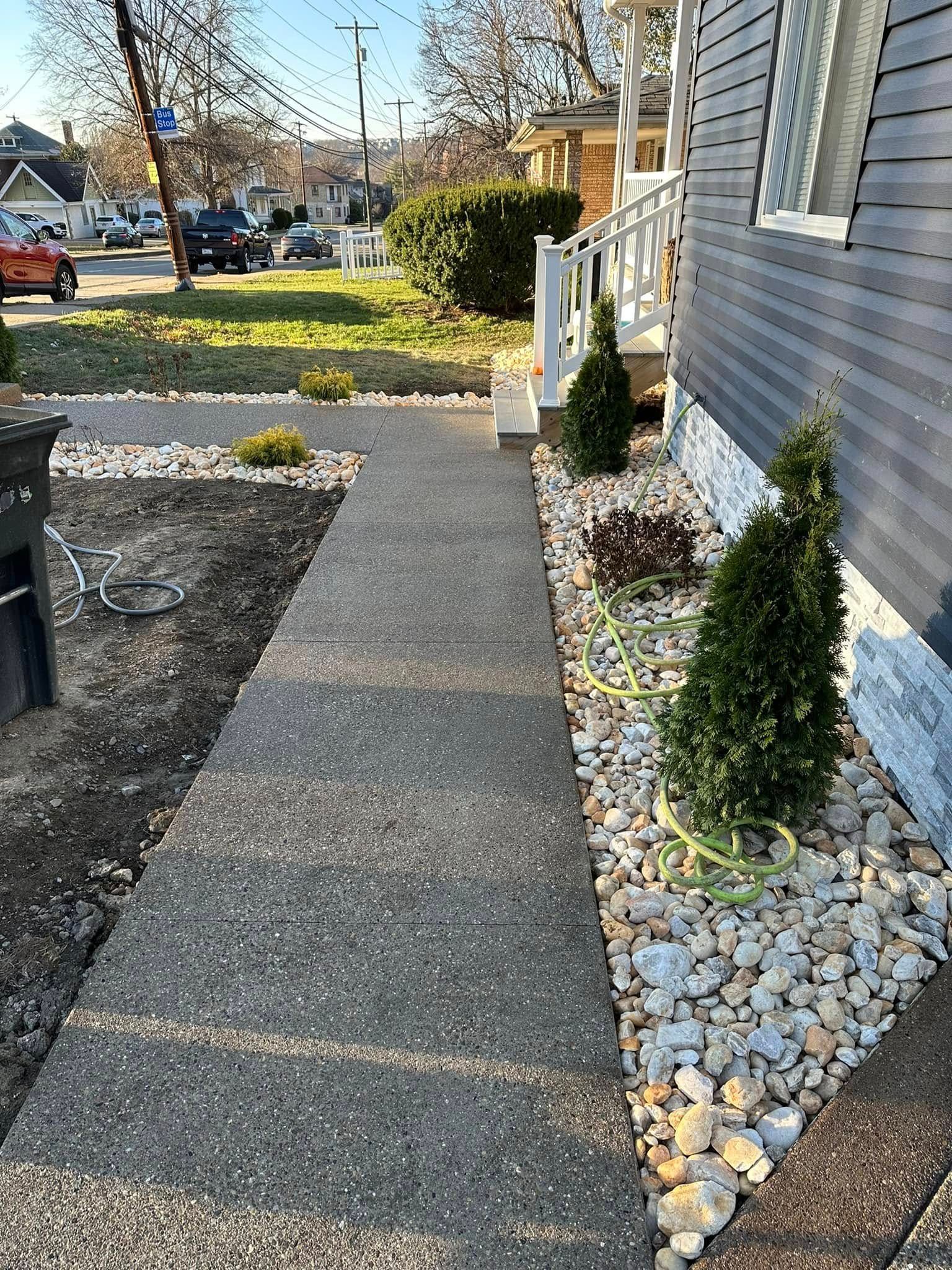 Concrete walkway beside a gray house, bordered by white rocks and small evergreen shrubs on a sunny day