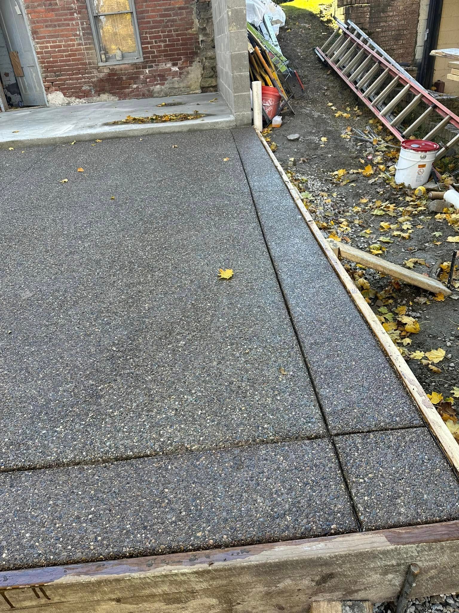 Concrete driveway beside brick wall and ladder, with fallen leaves and a garden border.