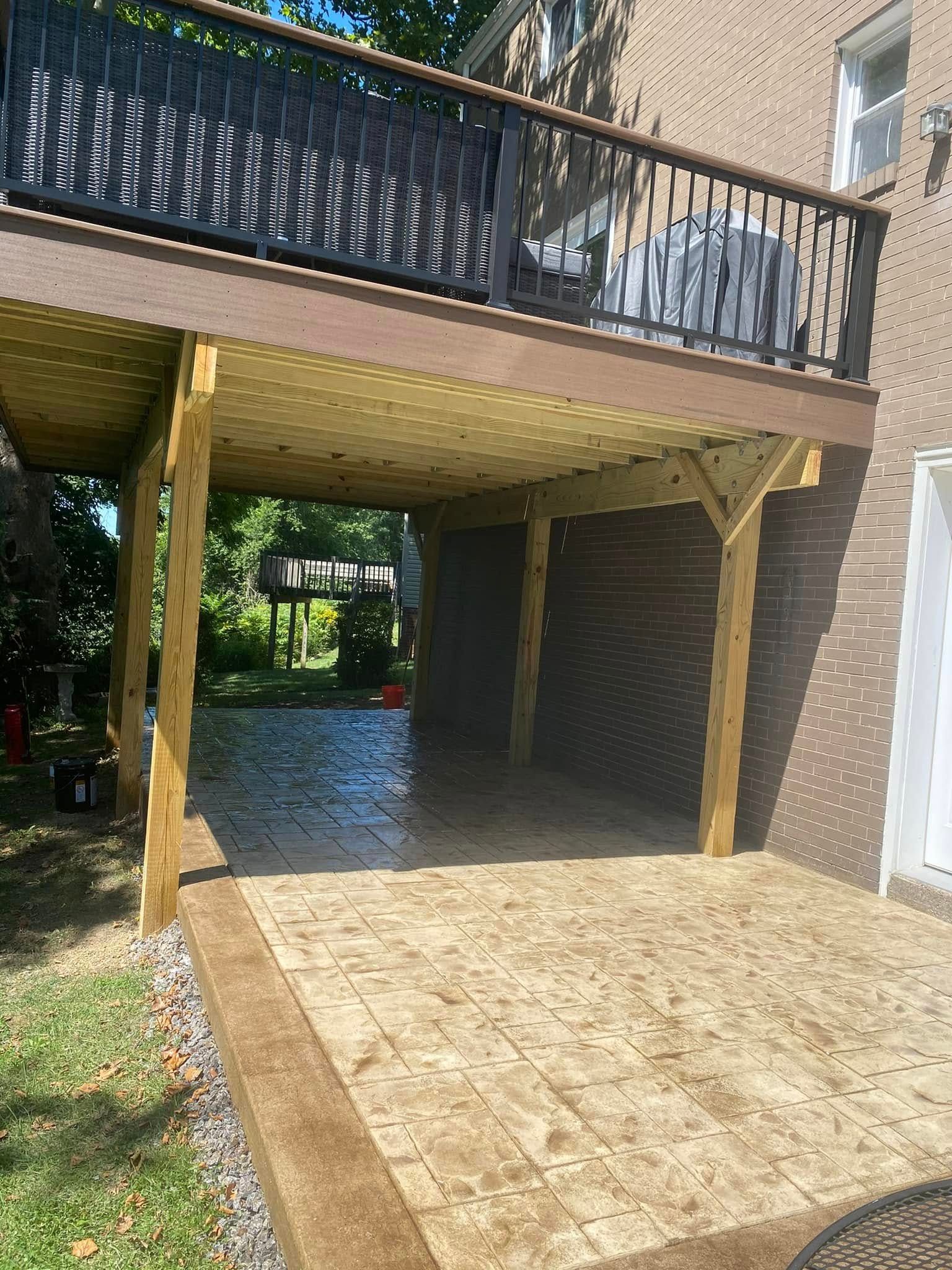 Covered wooden patio under a raised deck beside a brick house, with a paved walkway and shaded area below.