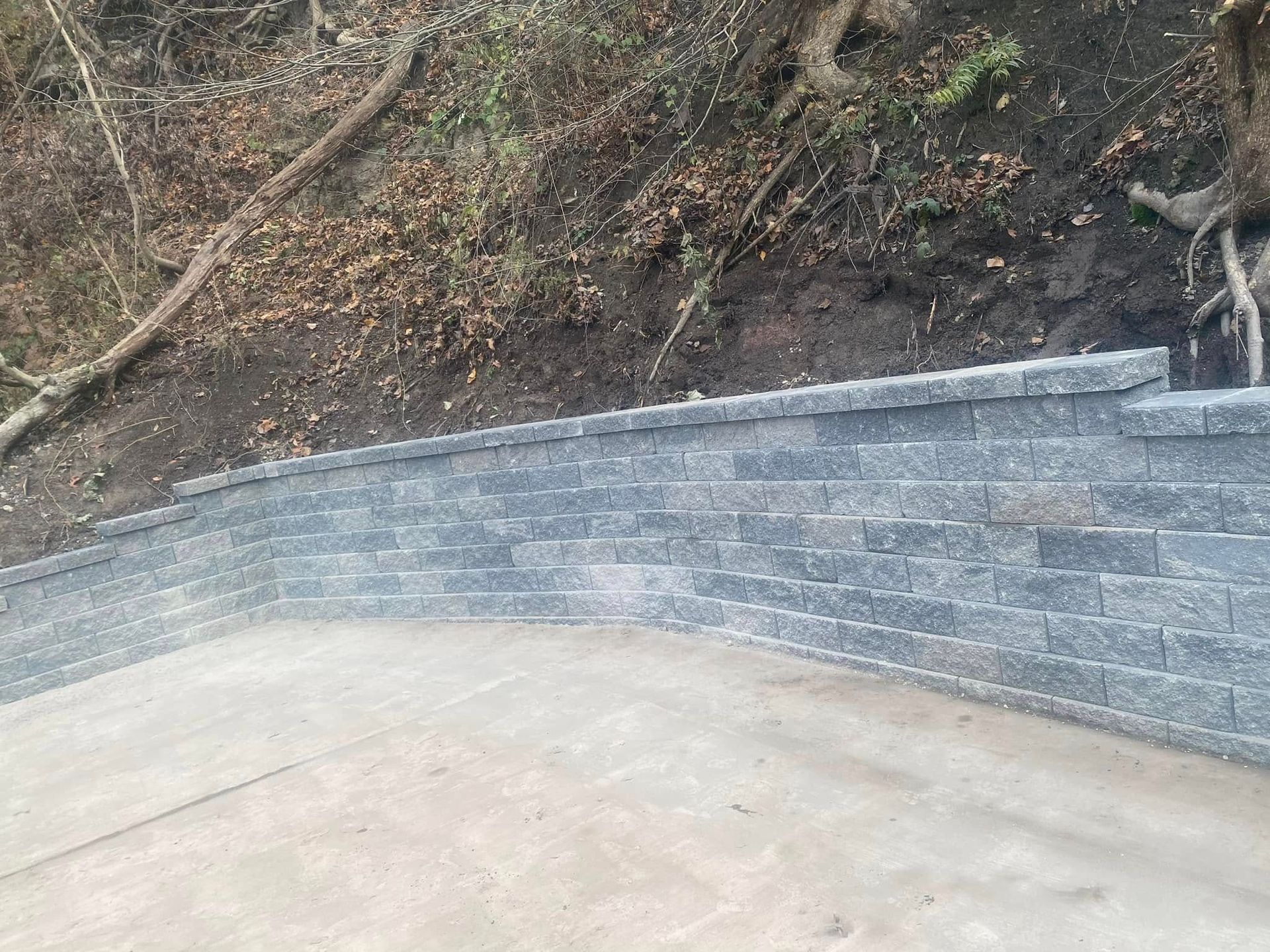 Curved gray stone retaining wall beside a concrete patio and dirt hillside with exposed roots