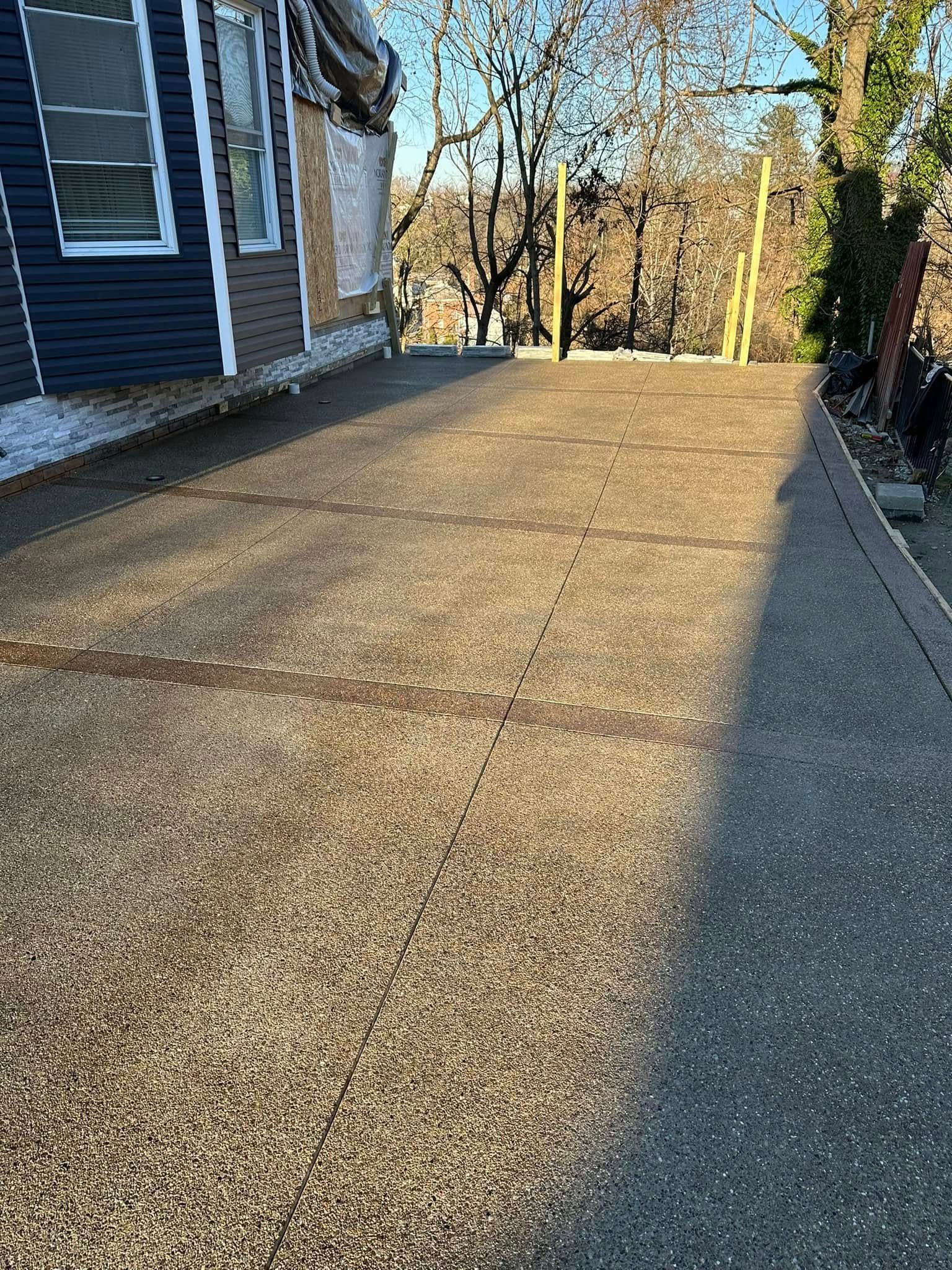 Concrete walkway beside a house, leading toward trees in a sunny yard.
