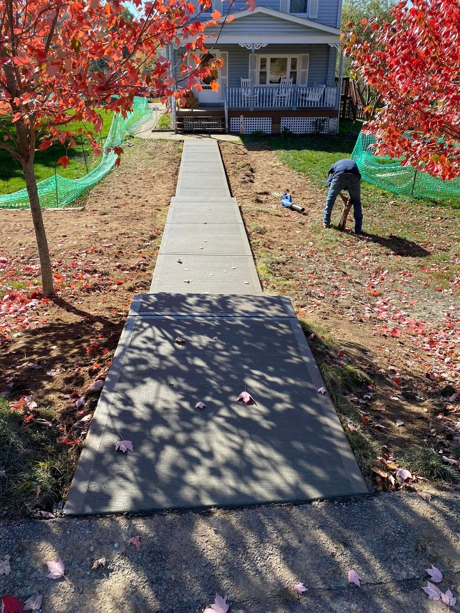Concrete walkway leading to a porch, framed by red autumn trees and fallen leaves.