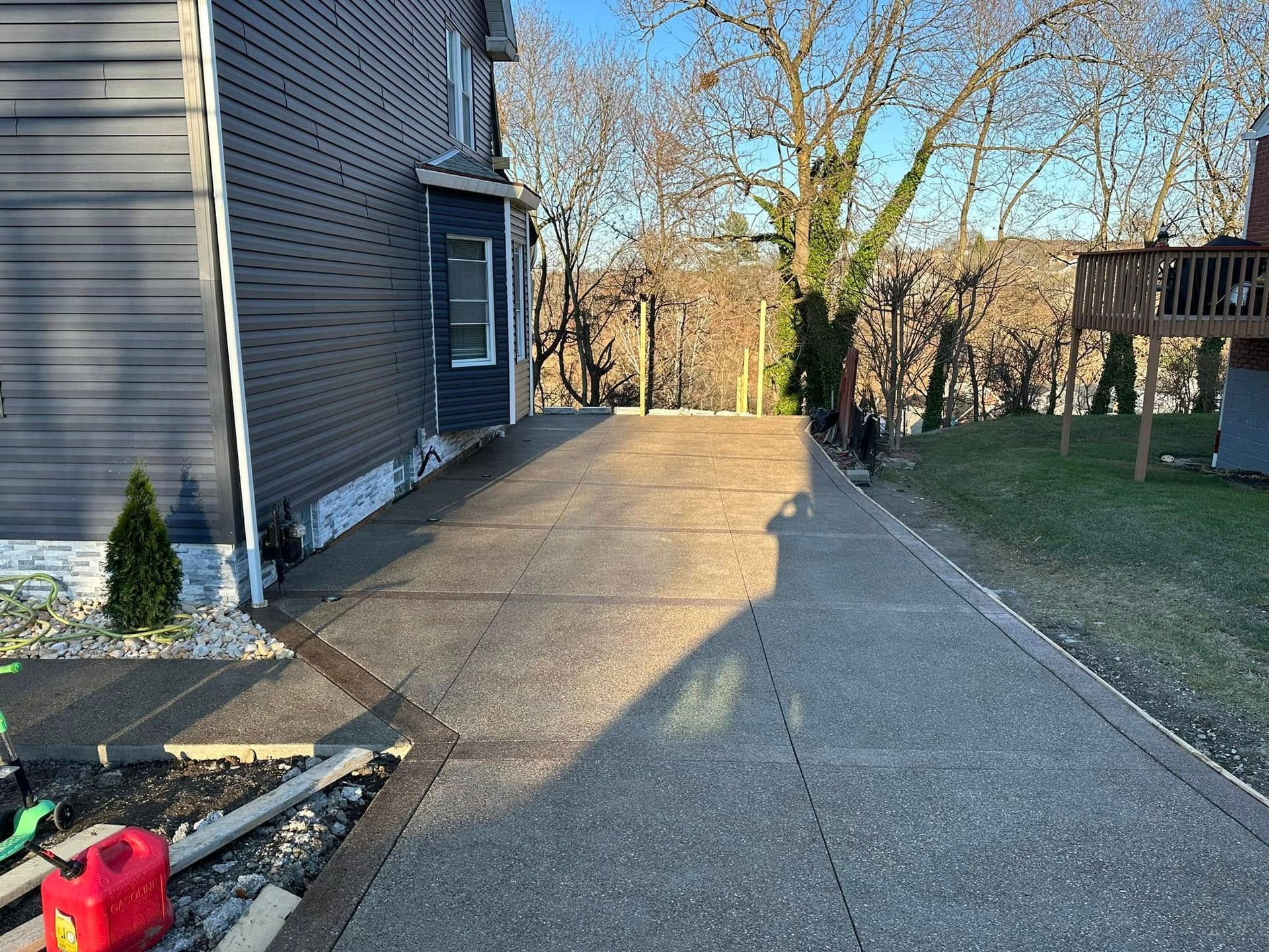 Side driveway beside a gray house, with concrete path and long shadows in a sunny yard.