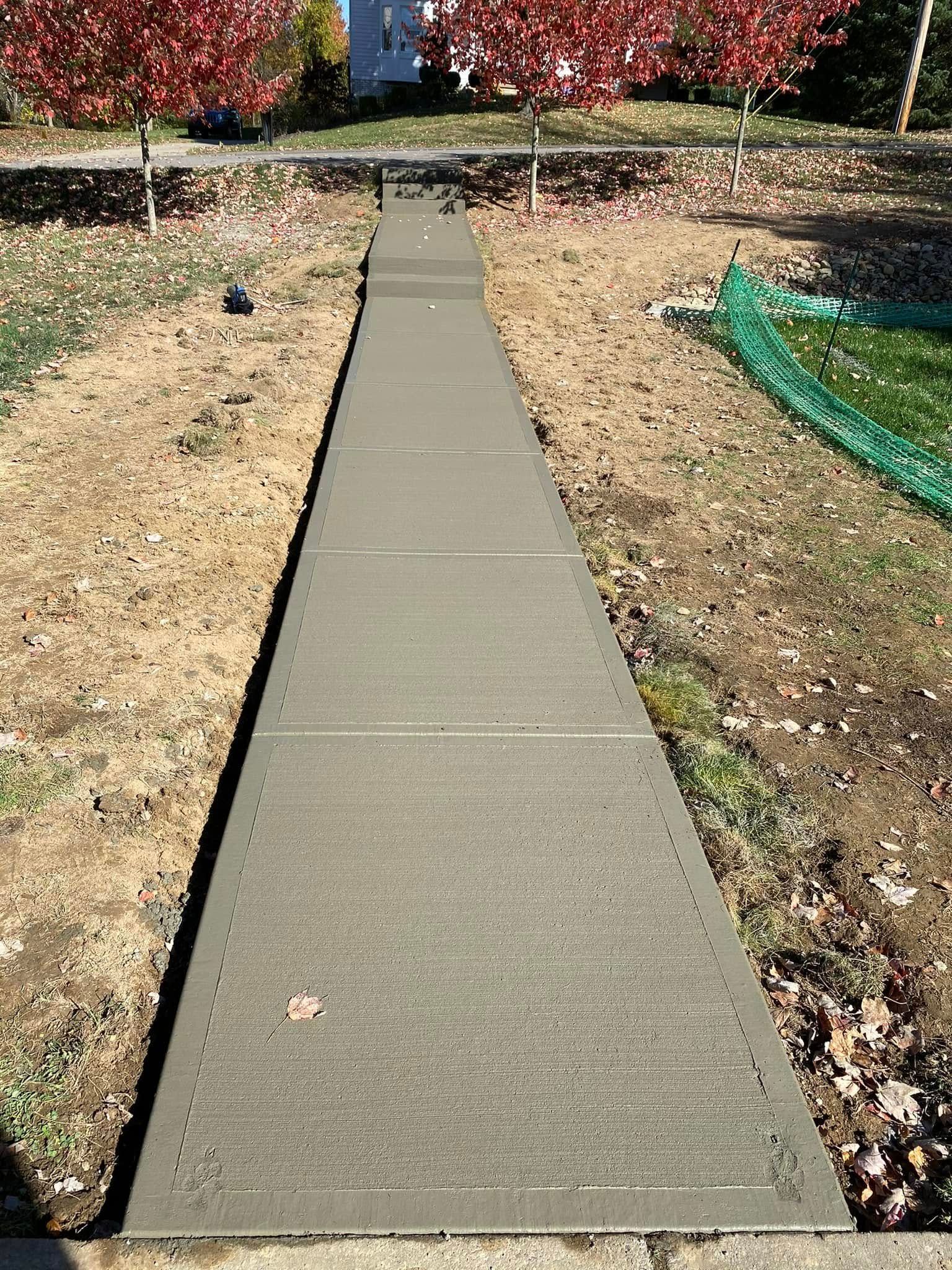 Concrete sidewalk through a dry yard with red autumn trees in the background