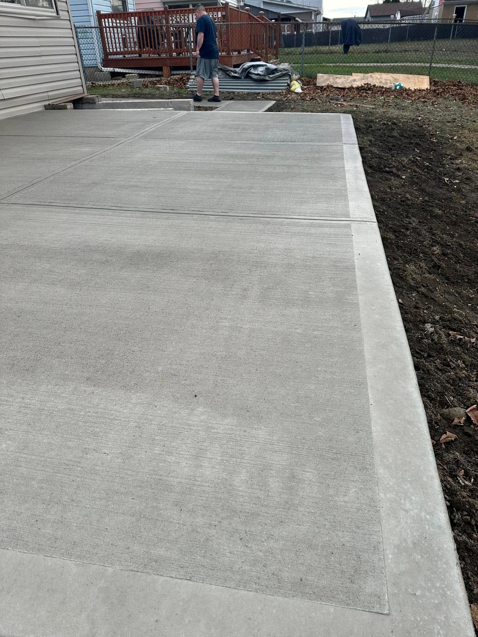 Concrete walkway beside a landscaped bed, with a person near a wooden deck in the background.