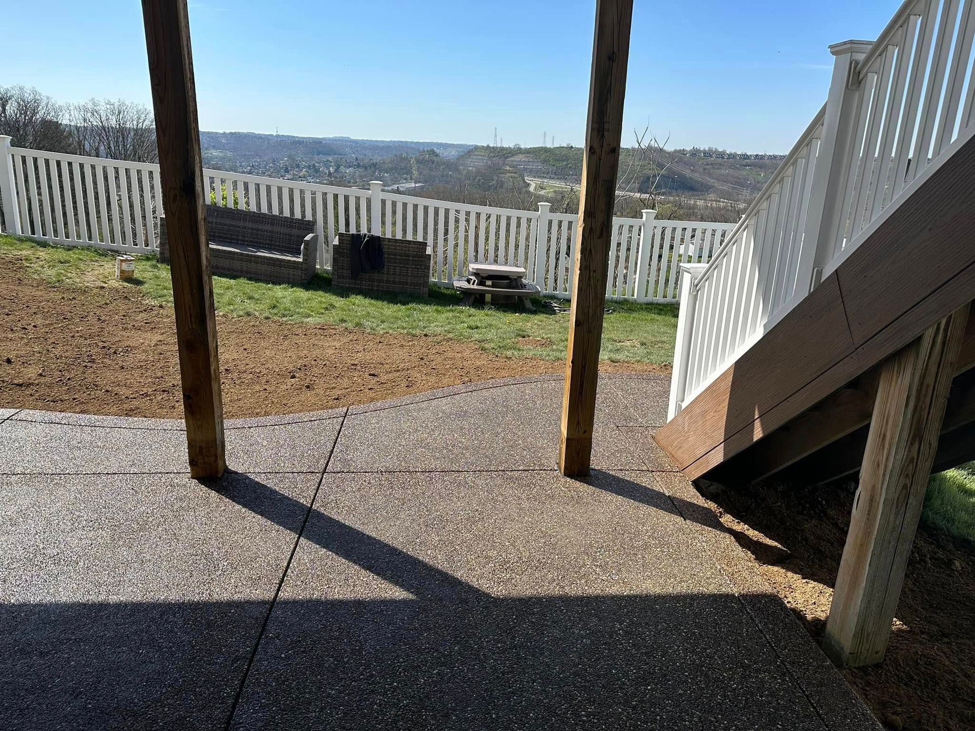 Backyard patio under a raised deck with white railings, gravel yard, and distant hills beyond a fence