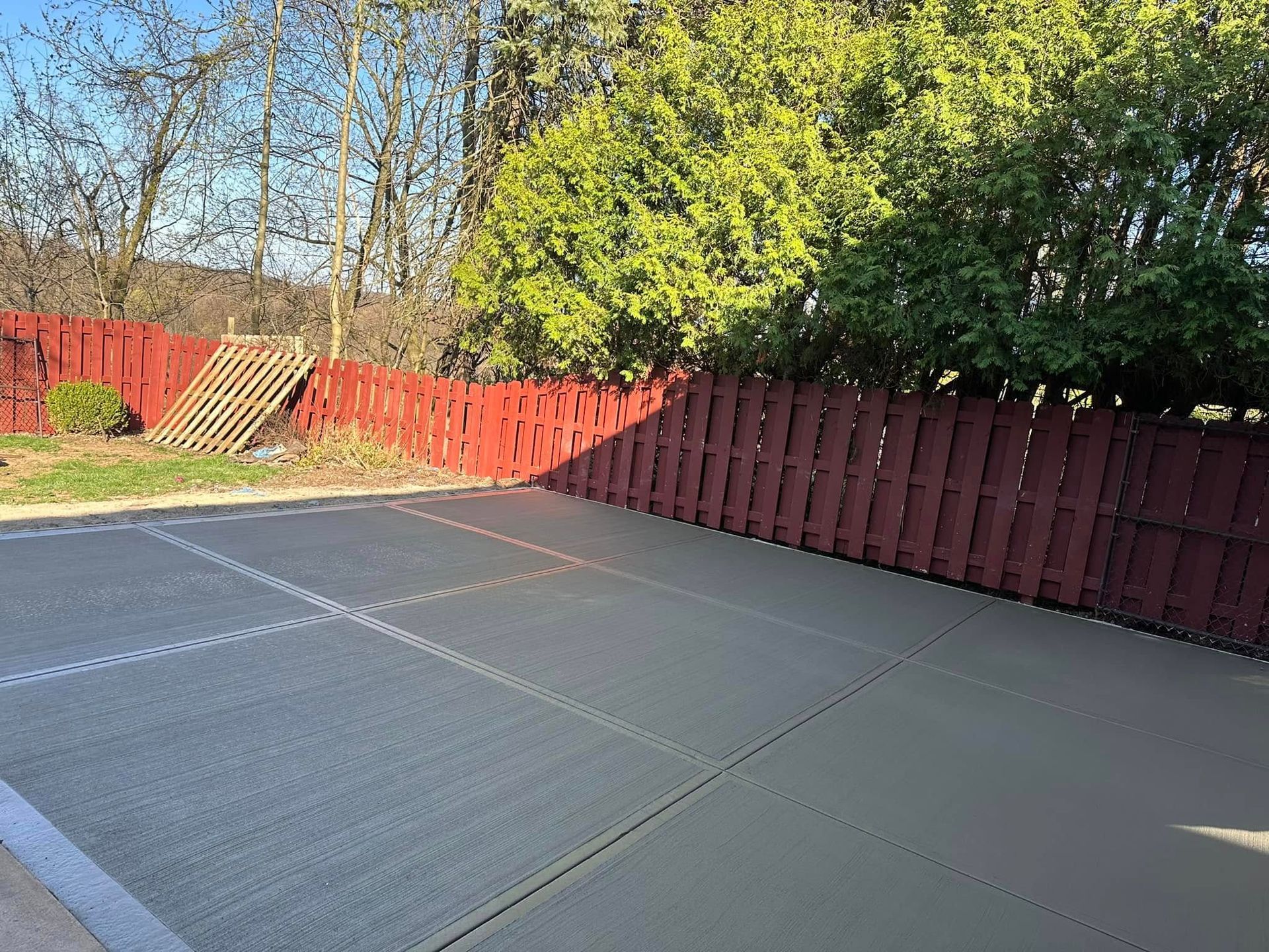 Empty backyard patio with gray mats, red wooden fence, and trees in bright sunlight