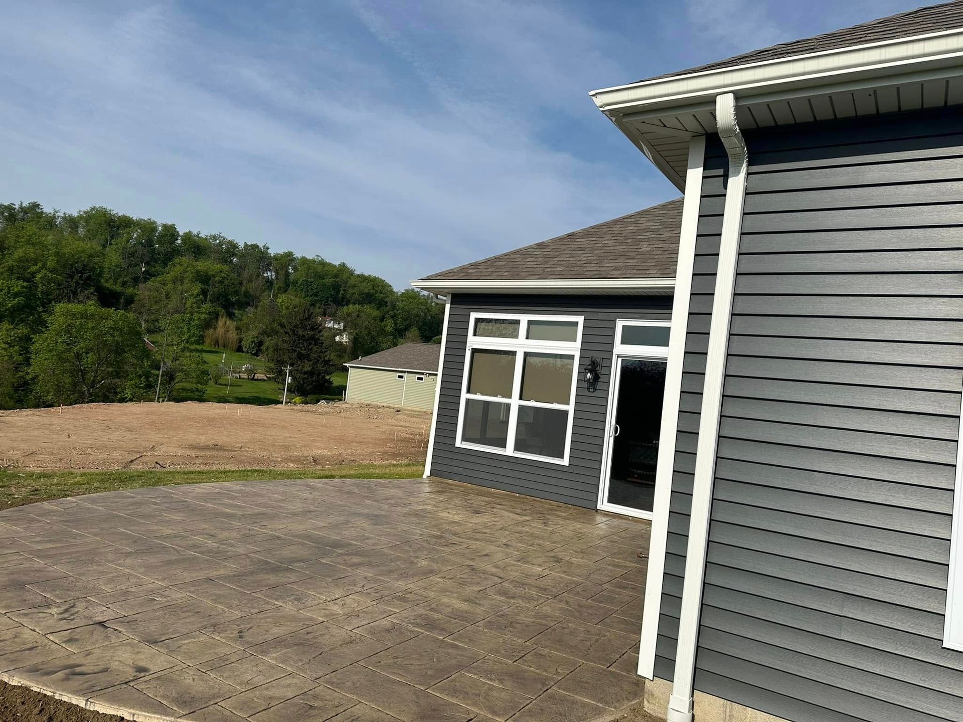 Back patio beside a gray house, with a paved area and grassy yard under a blue sky.
