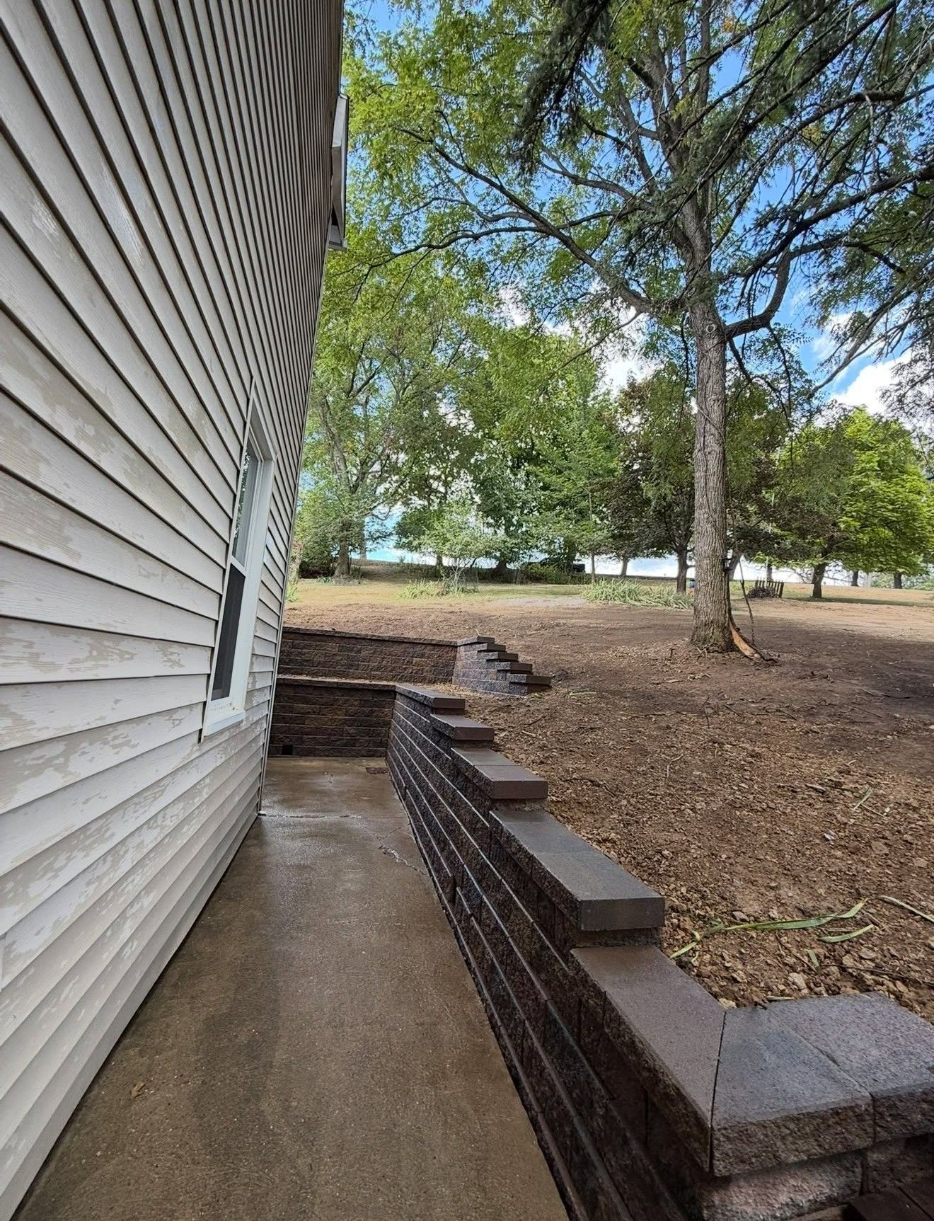 Narrow concrete walkway beside a white house, bordered by a low brick wall and shaded trees.
