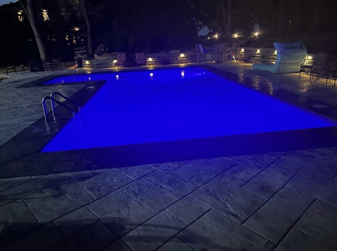 Nighttime outdoor pool glowing blue, surrounded by lounge chairs and patio lights.