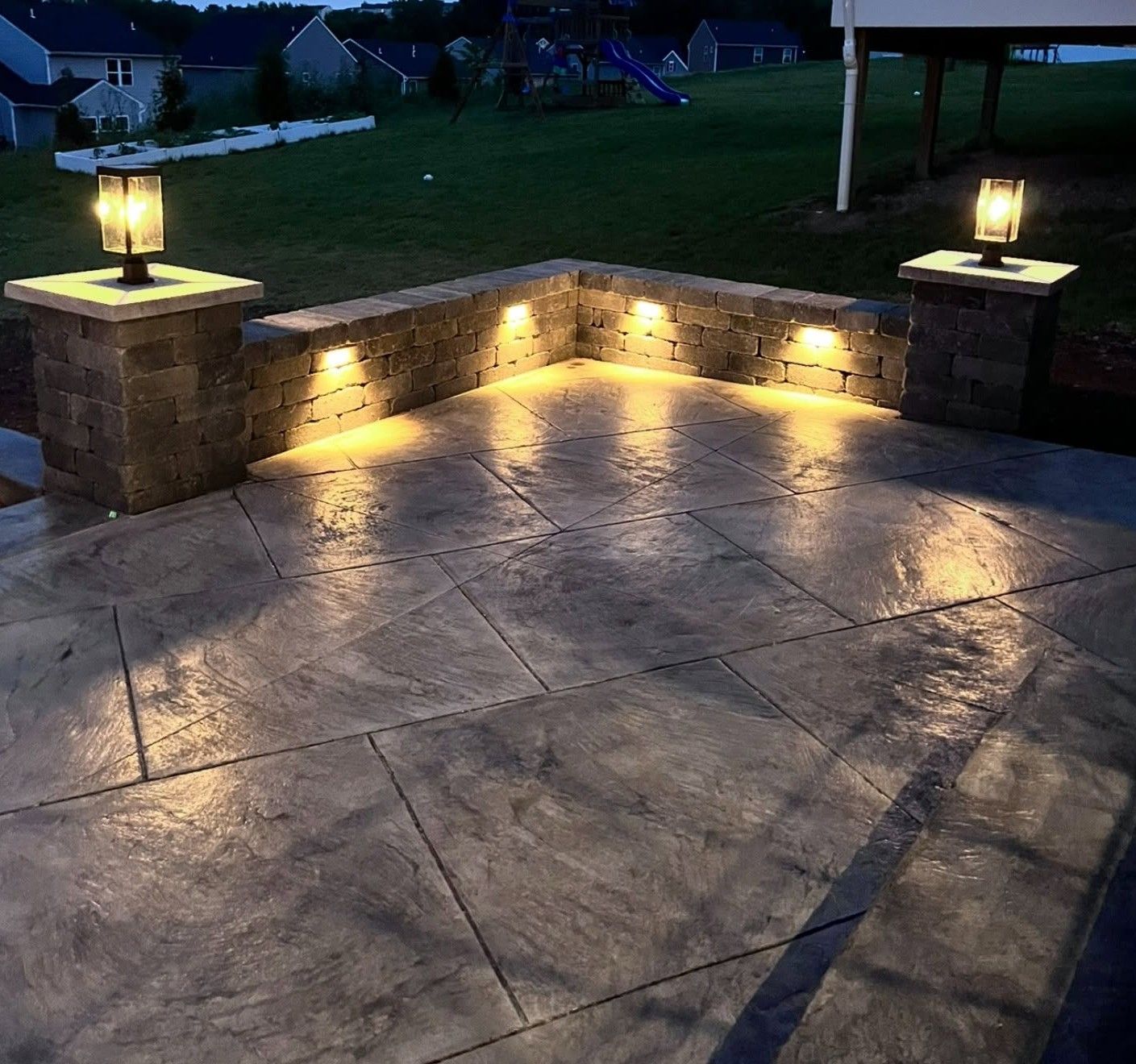 Stone patio at dusk with lit lanterns on two pillars and warm accent lights along the retaining wall.