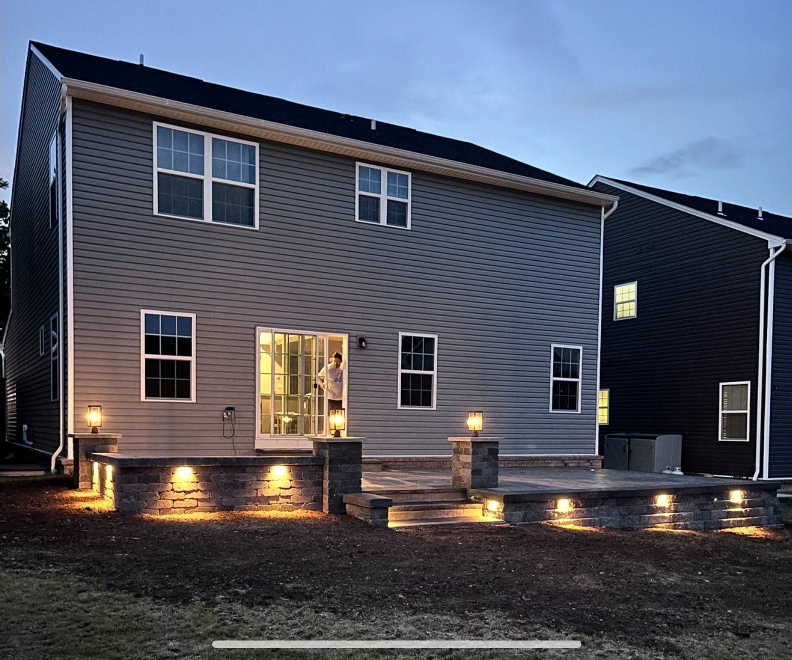 Backyard of a modern two-story house at dusk, with warm exterior lights illuminating the patio and lawn.