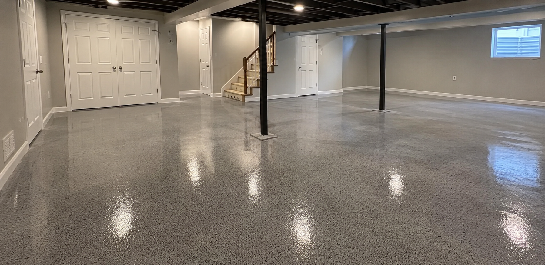 Empty basement room with polished concrete floor, white walls, exposed beams, and a staircase