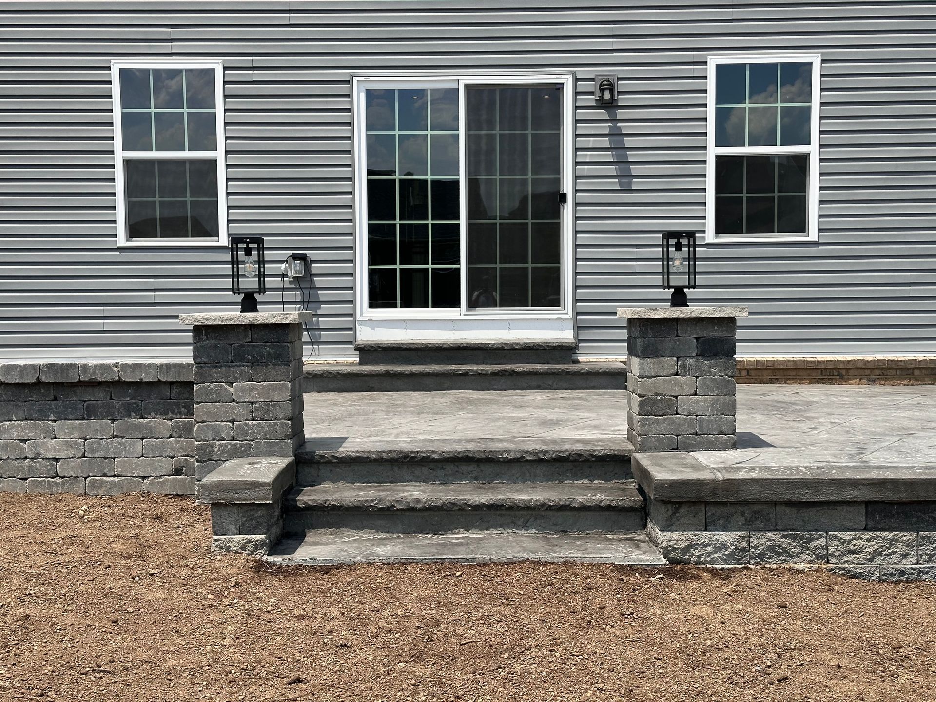 Gray house back steps with white-trimmed sliding glass door, two windows, and stone pillars beside a gravel yard