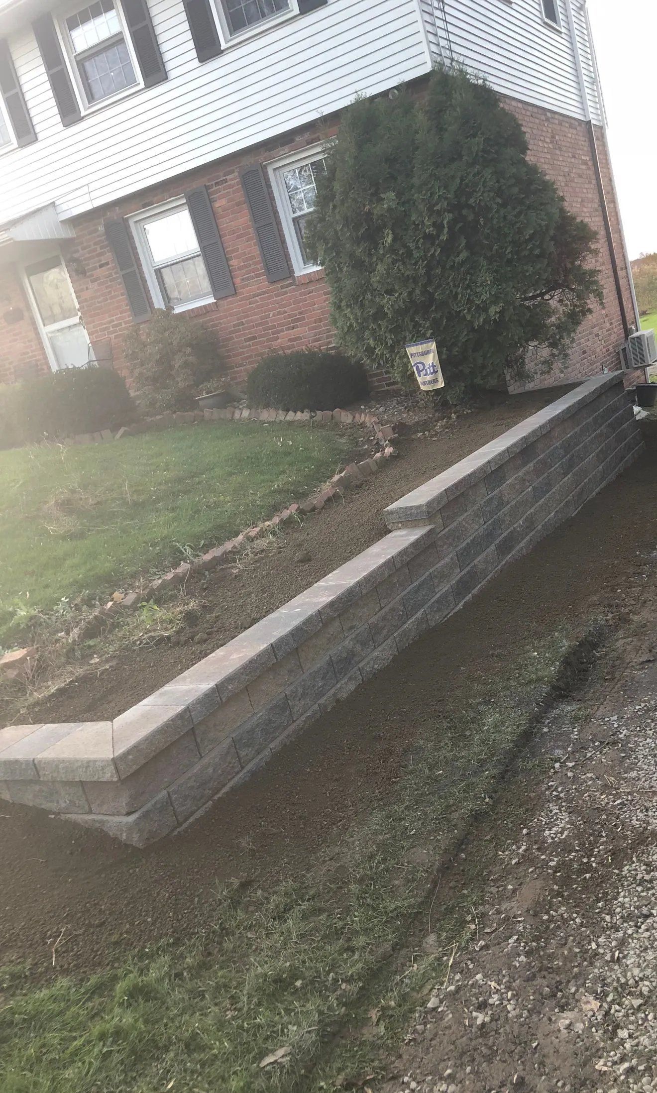 Brick townhouse with a sloped front yard, low stone retaining wall, and shrubs along the walkway.