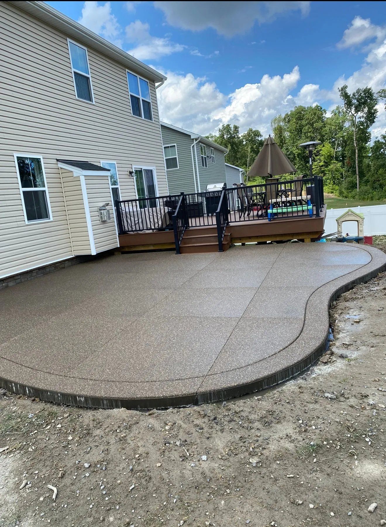 Back patio with curved stamped concrete, black railing, and tan siding beside a house under a blue sky