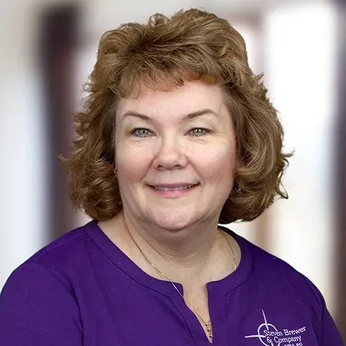 A person with shoulder-length wavy brown hair smiles at the camera, wearing a purple shirt with a small white logo.