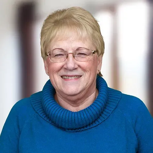 A smiling person wearing glasses and a blue cowl-neck sweater against a blurred background.