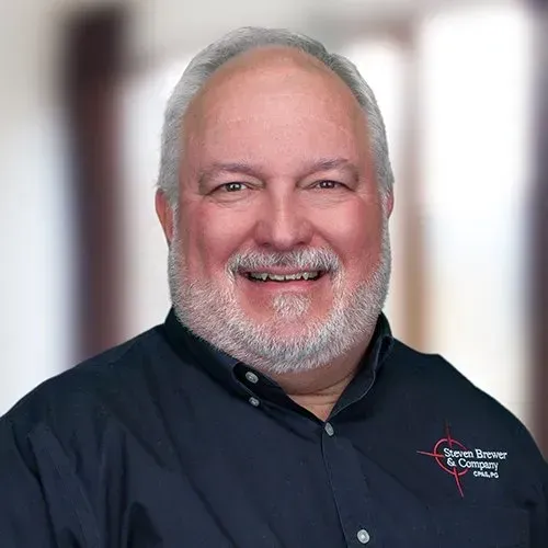 A smiling individual in a dark blue shirt with a small company logo on the chest, set against a blurred background.