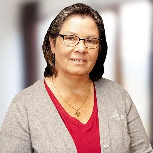 A woman with glasses and dark hair wearing a grey cardigan over a red shirt, smiling slightly against a blurred background.