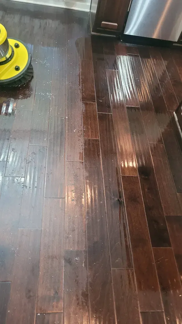 Wet, dark hardwood floor being cleaned by a yellow and black floor polisher in a kitchen.