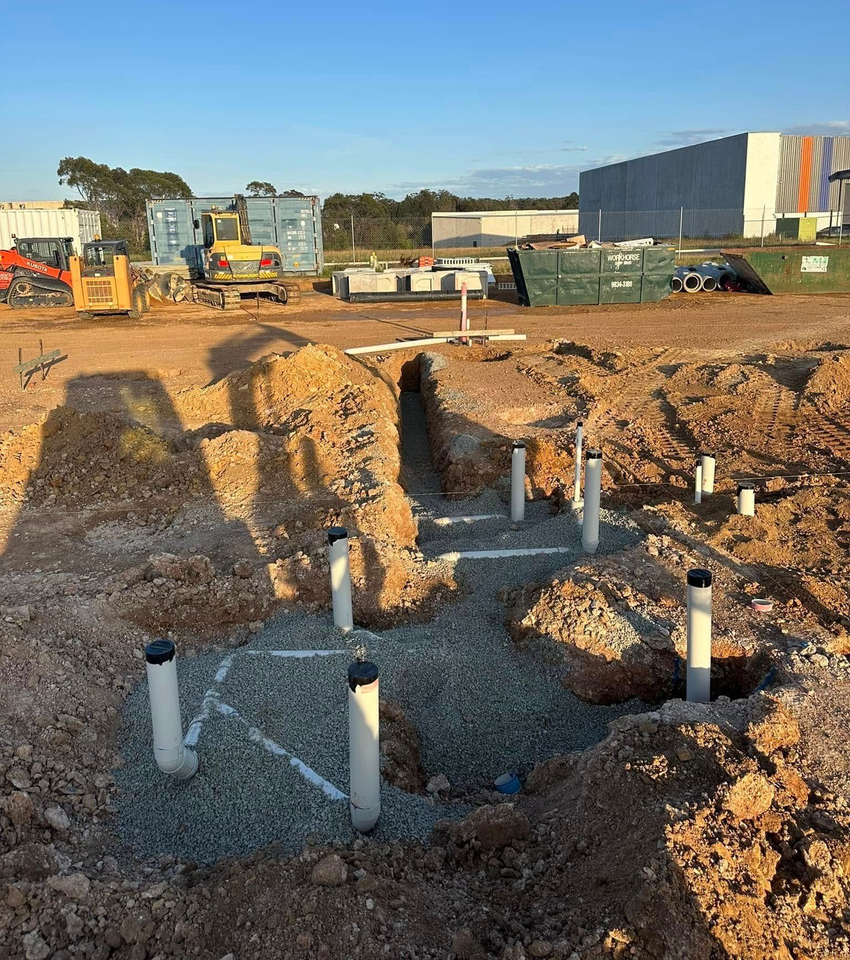 Construction site with dug trenches, exposed pipes, and heavy machinery under a clear sky