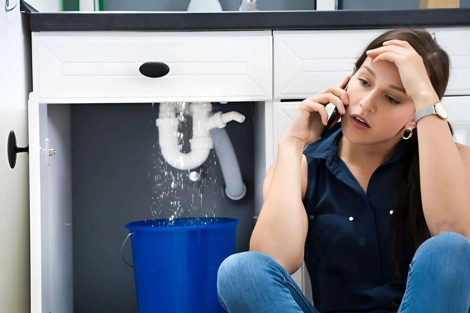 Woman sitting under a leaking sink, talking on the phone beside a blue bucket catching water