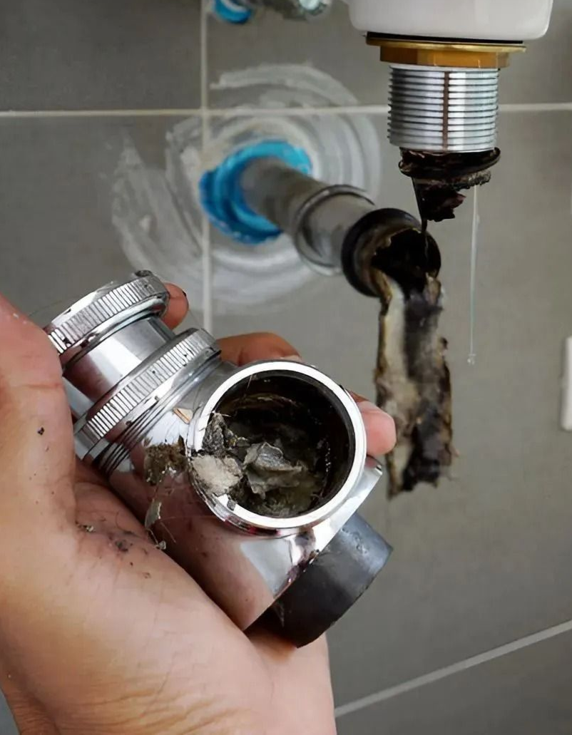Hand holding a clogged metal plumbing drain trap beneath a sink, with dark buildup inside and a leaking pipe above